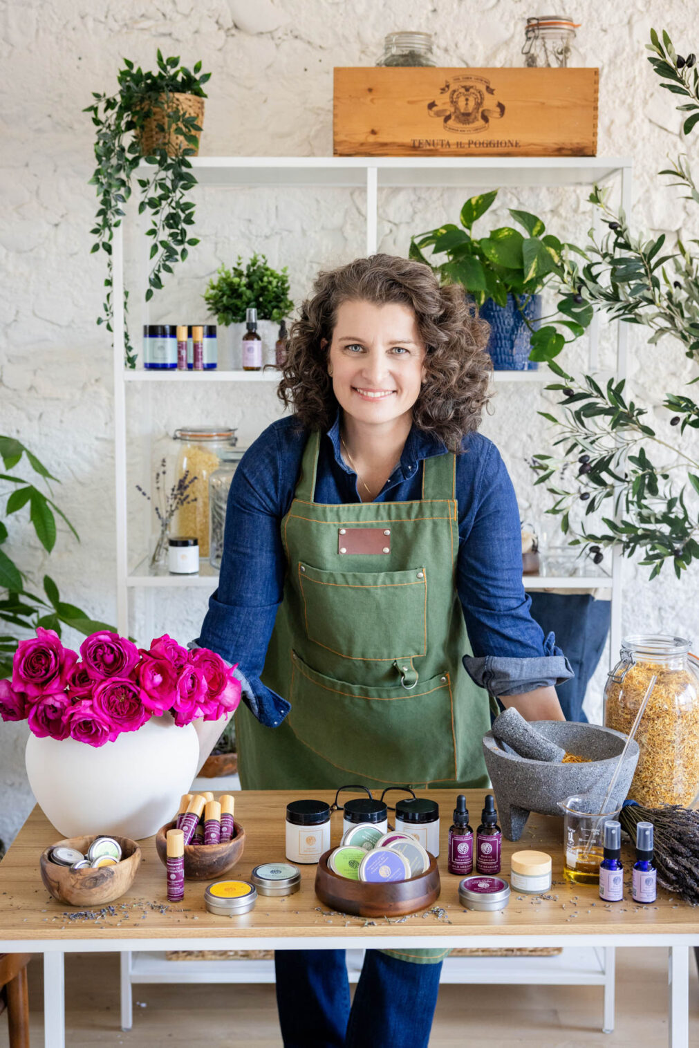 brand photo of a woman in an apron behind a desk with her skin care products and in front of a shelf holding plants, herbs and other skin care products