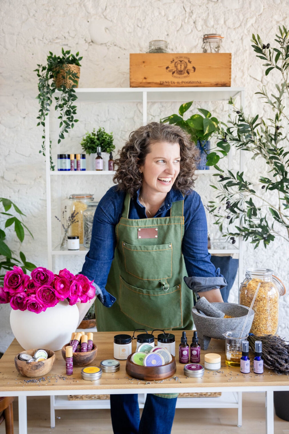 woman in green apron leans on a table with flowers and skincare products on it and looks to the side and laughs