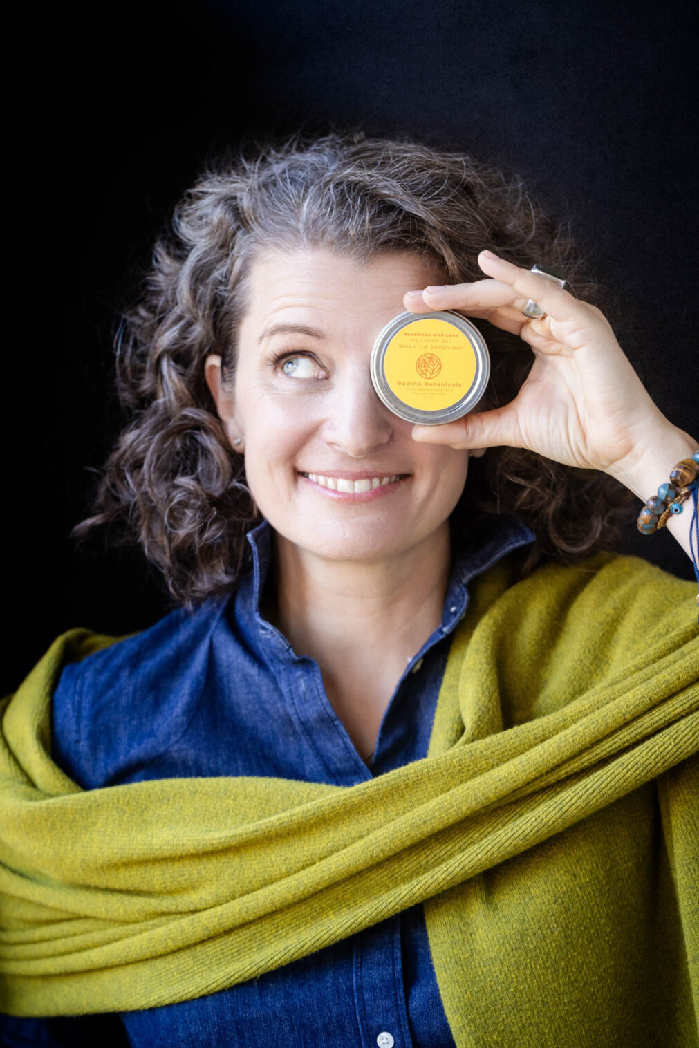 woman in a green wrap holds a round hand balm tin up to her eye